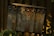 A row of old, rusted metal mailboxes is mounted against a wooden fence, partially obscured by surrounding leaves and plants. The mailboxes display faded numbers and are set in a dimly lit, outdoor area with a natural, rustic appearance.
