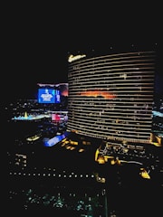 A nighttime cityscape featuring a tall, illuminated hotel building with horizontal light patterns. Adjacent buildings and areas are adorned with bright neon lights and digital screens displaying vibrant colors, contributing to the lively urban atmosphere.