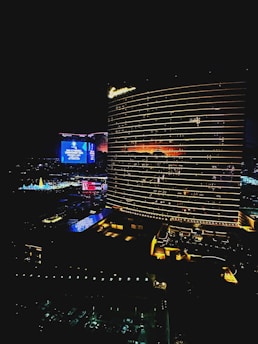 A nighttime cityscape featuring a tall, illuminated hotel building with horizontal light patterns. Adjacent buildings and areas are adorned with bright neon lights and digital screens displaying vibrant colors, contributing to the lively urban atmosphere.