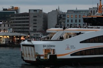 A ferry named 'ŞH-Göksu' is docked at a port in Istanbul, with people visible on board. The vessel is painted white with orange accents and the cityscape features several modern buildings in the background, including one with illuminated windows. The sky appears overcast, indicating a cloudy day.