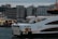 A ferry named 'ŞH-Göksu' is docked at a port in Istanbul, with people visible on board. The vessel is painted white with orange accents and the cityscape features several modern buildings in the background, including one with illuminated windows. The sky appears overcast, indicating a cloudy day.