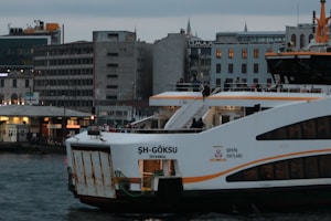 A ferry named 'ŞH-Göksu' is docked at a port in Istanbul, with people visible on board. The vessel is painted white with orange accents and the cityscape features several modern buildings in the background, including one with illuminated windows. The sky appears overcast, indicating a cloudy day.
