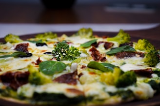 A close-up view of a pizza topped with broccoli, spinach leaves, olives, sundried tomatoes, and melted cheese. The pizza has a vibrant mix of green and red toppings, with a garnish of fresh parsley at the center.