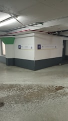 A concrete parking garage corner with visible structural elements, featuring directional exit signs on the walls. The environment appears slightly worn with visible moisture on the ground and overhead lighting. Pipes and conduits are attached to the ceiling.