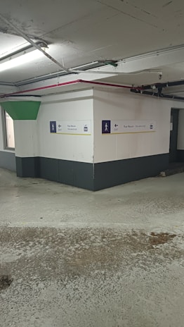 A concrete parking garage corner with visible structural elements, featuring directional exit signs on the walls. The environment appears slightly worn with visible moisture on the ground and overhead lighting. Pipes and conduits are attached to the ceiling.