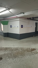 A concrete parking garage corner with visible structural elements, featuring directional exit signs on the walls. The environment appears slightly worn with visible moisture on the ground and overhead lighting. Pipes and conduits are attached to the ceiling.
