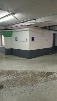 A concrete parking garage corner with visible structural elements, featuring directional exit signs on the walls. The environment appears slightly worn with visible moisture on the ground and overhead lighting. Pipes and conduits are attached to the ceiling.