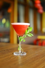 A cocktail glass filled with a bright red beverage sits on a wooden table. The drink is garnished with fresh green mint leaves wrapped around the stem of the glass. The background is softly blurred, featuring warm tones and ambient lighting.