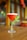 A cocktail glass filled with a bright red beverage sits on a wooden table. The drink is garnished with fresh green mint leaves wrapped around the stem of the glass. The background is softly blurred, featuring warm tones and ambient lighting.