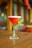 A cocktail glass filled with a bright red beverage sits on a wooden table. The drink is garnished with fresh green mint leaves wrapped around the stem of the glass. The background is softly blurred, featuring warm tones and ambient lighting.