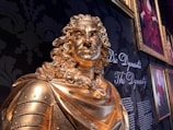 A golden statue of a historical figure with detailed facial features and curly hair is prominently displayed. The background features a dark floral pattern and a gallery wall with framed portraits and the words 'Die Dynastie' and 'The Dynasty' written in elegant script.