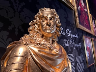A golden statue of a historical figure with detailed facial features and curly hair is prominently displayed. The background features a dark floral pattern and a gallery wall with framed portraits and the words 'Die Dynastie' and 'The Dynasty' written in elegant script.