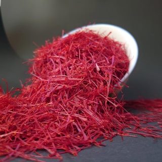Bright red saffron threads are spilling out of a tilted white bowl onto a dark surface. The fine, delicate threads create a vivid contrast against the dark background.