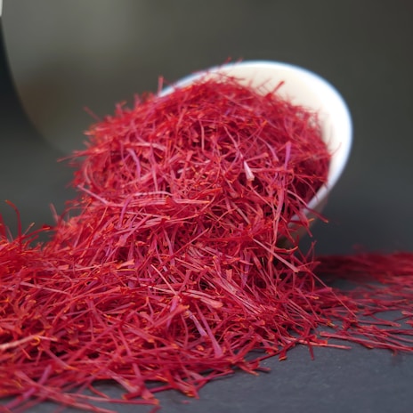 Bright red saffron threads are spilling out of a tilted white bowl onto a dark surface. The fine, delicate threads create a vivid contrast against the dark background.