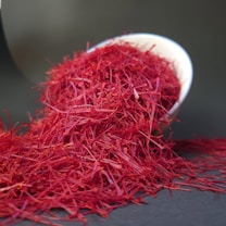 Bright red saffron threads are spilling out of a tilted white bowl onto a dark surface. The fine, delicate threads create a vivid contrast against the dark background.