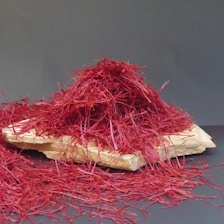 A pile of vibrant red saffron threads is arranged on a flat, light-colored wooden surface. The threads are thin, intricate, and densely packed, creating an almost mound-like appearance. The background is a plain, muted gray, which contrasts with the bright red and helps the saffron stand out prominently.