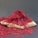 A pile of vibrant red saffron threads is arranged on a flat, light-colored wooden surface. The threads are thin, intricate, and densely packed, creating an almost mound-like appearance. The background is a plain, muted gray, which contrasts with the bright red and helps the saffron stand out prominently.