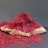 A pile of vibrant red saffron threads is arranged on a flat, light-colored wooden surface. The threads are thin, intricate, and densely packed, creating an almost mound-like appearance. The background is a plain, muted gray, which contrasts with the bright red and helps the saffron stand out prominently.