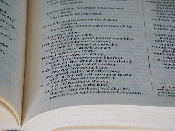 An open book displaying text on two pages. The content appears to be printed in a poetic format, with verse numbers on the left side of each line. The pages are light-colored, and the text is in a black serif font. The book is opened wide, revealing the curvature of the spine.