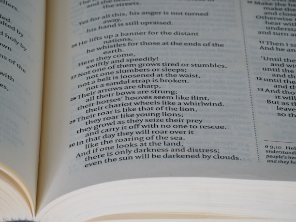 An open book displaying text on two pages. The content appears to be printed in a poetic format, with verse numbers on the left side of each line. The pages are light-colored, and the text is in a black serif font. The book is opened wide, revealing the curvature of the spine.