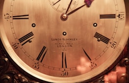 An antique clock face features Roman numerals, ornate hands, and the inscription 'Lund & Blockley, to the Queen, 42 Pall Mall, London.' The clock has a polished brass finish and intricate wood detailing around the edges, suggesting a classic and luxurious design.