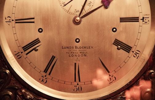 An antique clock face features Roman numerals, ornate hands, and the inscription 'Lund & Blockley, to the Queen, 42 Pall Mall, London.' The clock has a polished brass finish and intricate wood detailing around the edges, suggesting a classic and luxurious design.