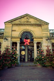 An entrance of a traditional market building adorned with festive decorations. Two decorated Christmas trees with red and gold ornaments flank the entrance. A large red bow is prominently displayed above the entry with a wreath around the circular window. The building is made of stone with columns, and a cobblestone path leads to the doorway.