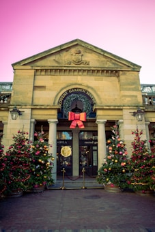 An entrance of a traditional market building adorned with festive decorations. Two decorated Christmas trees with red and gold ornaments flank the entrance. A large red bow is prominently displayed above the entry with a wreath around the circular window. The building is made of stone with columns, and a cobblestone path leads to the doorway.