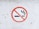 A circular no smoking sign on a white, slightly textured surface. The sign features a cigarette with smoke rising, crossed out by a red circle with a diagonal line.