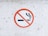 A circular no smoking sign on a white, slightly textured surface. The sign features a cigarette with smoke rising, crossed out by a red circle with a diagonal line.