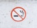 A circular no smoking sign on a white, slightly textured surface. The sign features a cigarette with smoke rising, crossed out by a red circle with a diagonal line.