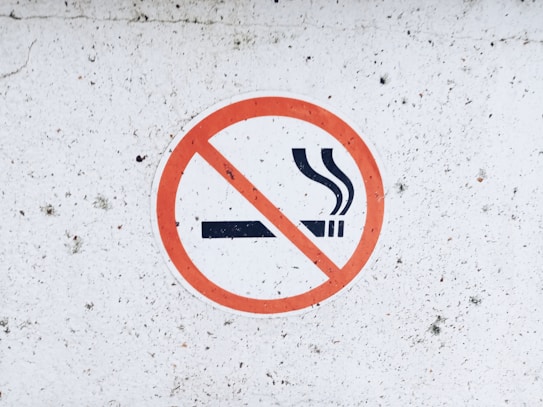 A circular no smoking sign on a white, slightly textured surface. The sign features a cigarette with smoke rising, crossed out by a red circle with a diagonal line.