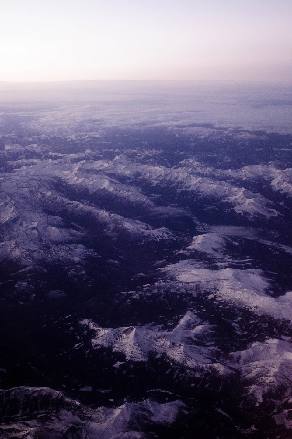 The image shows an aerial view of a mountainous landscape, captured from the perspective of an airplane window. The mountains are blanketed in snow and exhibit a range of colors, including shades of blue, green, and brown. The sky above is a light blue color with wispy clouds scattered across it. The image conveys a sense of vastness and grandeur as the viewer looks down upon this breathtaking natural scene from high above.