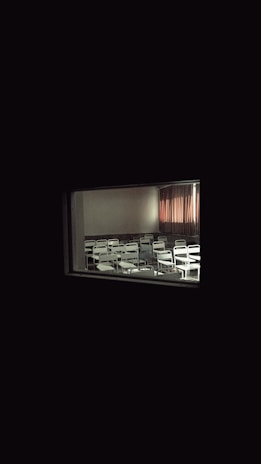 A dimly lit classroom with rows of empty white chairs and desks is visible through a small window. The room is mostly dark, with a curtain partly drawn to one side, allowing a faint glow of light to enter.