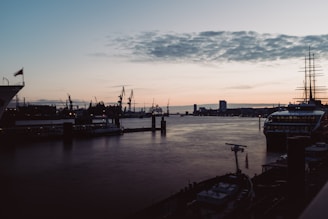 A calm harbor scene at sunset with moored cargo ships and cranes silhouetted against the sky.