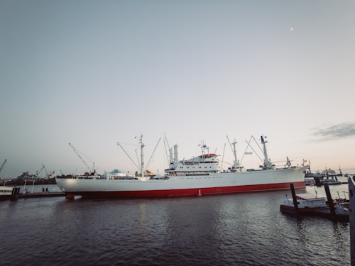 A large cargo ship docked at a harbor with cranes visible in the background. The ship is primarily white with a red bottom and features several masts and booms. The sky is clear with a hint of dusk or dawn, casting a serene ambiance over the scene.