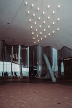A modern architectural interior with a reflective glass facade. The ceiling features a pattern of evenly spaced lights, and the floor is covered in a wooden texture. People are visible through the glass, walking along an outdoor terrace.