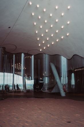 A modern architectural interior with a reflective glass facade. The ceiling features a pattern of evenly spaced lights, and the floor is covered in a wooden texture. People are visible through the glass, walking along an outdoor terrace.