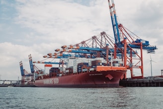 A large cargo ship is docked at a busy port, surrounded by multiple towering cranes painted in blue and orange. Shipping containers are stacked on the vessel, indicating active trade and commerce activity. The background features a partly cloudy sky and a wind turbine, suggesting sustainable energy use.