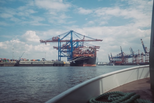 A bustling shipping port with a large cargo ship docked at a terminal. The scene includes several towering cranes used for loading and unloading containers, as well as stacks of colorful shipping containers along the dock. The sky is partly cloudy, casting a soft light over the port area. In the foreground, a boat's rail and a coiled rope are visible, indicating proximity to the water.