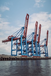 Large industrial cranes stand tall at a shipping port, with blue and red metal structures towering against a partly cloudy sky, reflecting a scene of bustling maritime commerce.