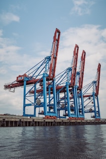 Large industrial cranes stand tall at a shipping port, with blue and red metal structures towering against a partly cloudy sky, reflecting a scene of bustling maritime commerce.