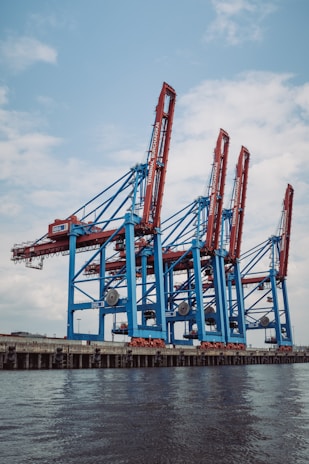 Large industrial cranes stand tall at a shipping port, with blue and red metal structures towering against a partly cloudy sky, reflecting a scene of bustling maritime commerce.