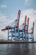 Large industrial cranes stand tall at a shipping port, with blue and red metal structures towering against a partly cloudy sky, reflecting a scene of bustling maritime commerce.