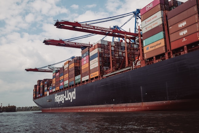 A large cargo ship is loaded with multiple shipping containers stacked high. Cranes labeled 'Terminal Burchardkai' are positioned above, suggesting the loading or unloading process is underway. The ship is sailing through a body of water with a cloudy sky overhead. The containers are colorful and bear various company logos.