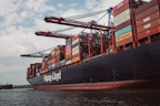 A large cargo ship is loaded with multiple shipping containers stacked high. Cranes labeled 'Terminal Burchardkai' are positioned above, suggesting the loading or unloading process is underway. The ship is sailing through a body of water with a cloudy sky overhead. The containers are colorful and bear various company logos.