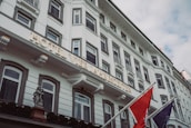 A large, elegant facade of a white hotel building featuring multiple rectangular windows with ornate frames. The hotel's name is displayed prominently in illuminated letters. Two flags are mounted on poles outside, and a decorative cherub statue is visible near the entrance.