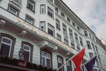 A large, elegant facade of a white hotel building featuring multiple rectangular windows with ornate frames. The hotel's name is displayed prominently in illuminated letters. Two flags are mounted on poles outside, and a decorative cherub statue is visible near the entrance.