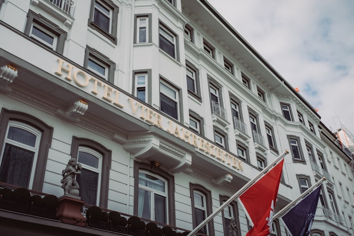 A large, elegant facade of a white hotel building featuring multiple rectangular windows with ornate frames. The hotel's name is displayed prominently in illuminated letters. Two flags are mounted on poles outside, and a decorative cherub statue is visible near the entrance.