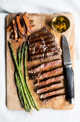 A wooden cutting board featuring a grilled steak sliced and arranged neatly, accompanied by a side of grilled asparagus and sweet potato fries. A small bowl of green herbs in oil is placed beside a sleek black-handled steak knife.